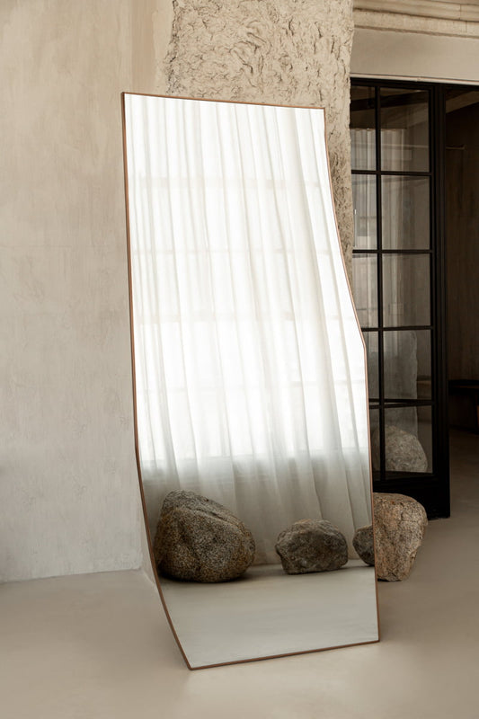 The Shard Mirror L reflects sheer white curtains and two large rocks in a minimalist, neutral-toned room with textured walls and a black-framed glass door, capturing modern elegance.