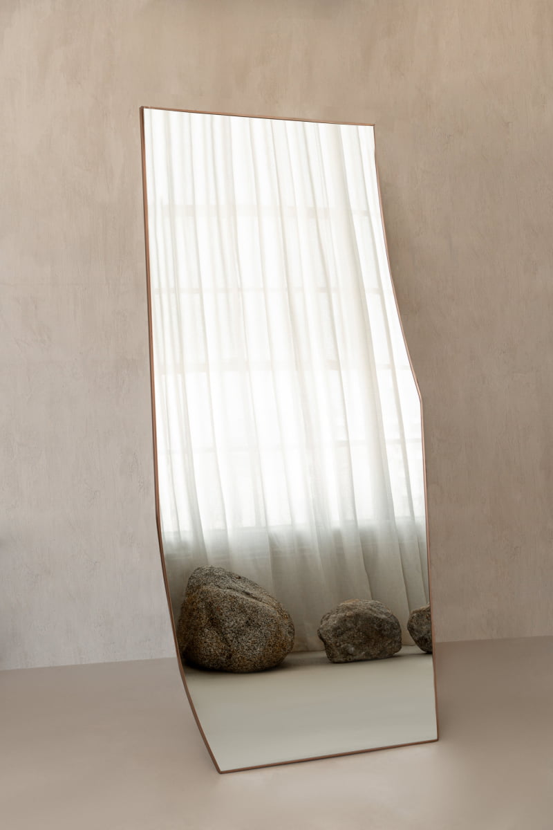 The Shard Mirror L, with its unique asymmetrical design, stands on the floor and reflects sheer white curtains and large rocks against a neutral wall, bringing modern elegance to the space.