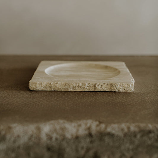 The Solara Charger is a square, beige stone tray with rough edges and a shallow circular indentation in the center, resting on a textured brown surface against a neutral background for a touch of natural elegance.