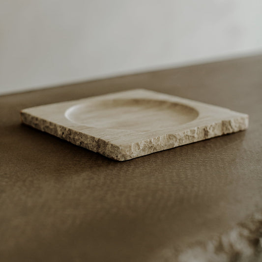 The Solara Charger, a square beige accessory with rough, unfinished edges, rests on a textured brown surface. It features a shallow, circular center indentation for a touch of natural elegance.
