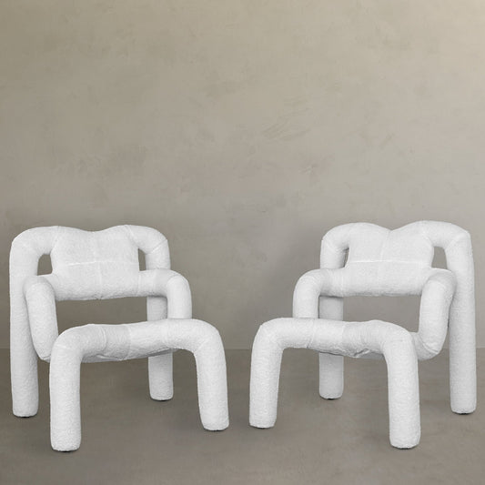 Four modern Spider Chair Boucle chairs with sculptural white neotenic geometry—curved legs and backs—are paired against a light wall and floor, creating a minimalist, contemporary setting and offering a unique seating experience.