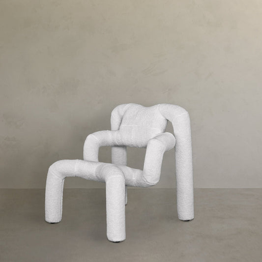 The Spider Chair Boucle features thick, rounded, white boucle-upholstered legs and armrests, its neotenic geometry and abstract modern design offering a unique sitting experience against a light grey textured wall.