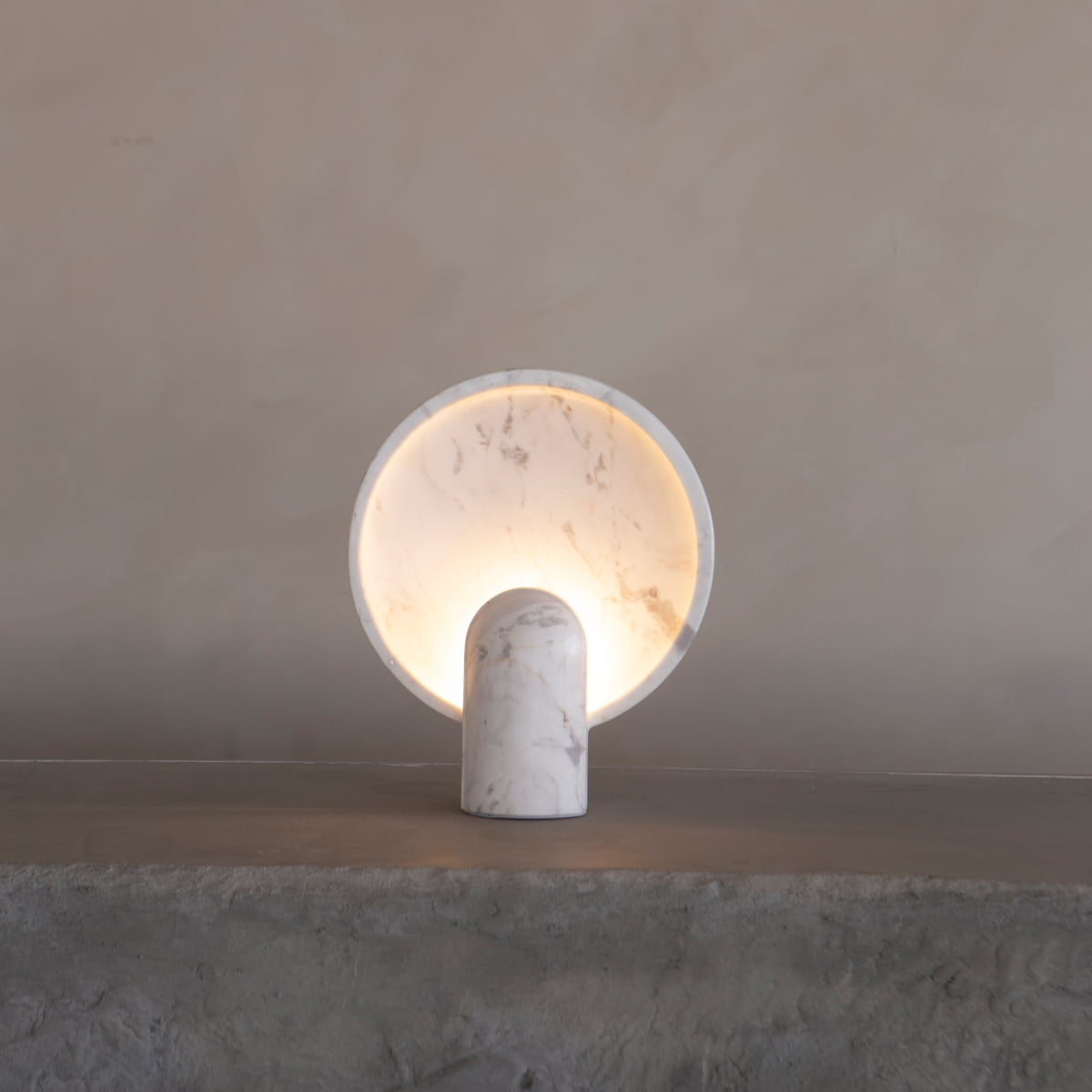 The Stella Lamp in Blanco Marble features a modern cylindrical base and round backplate, emitting a soft, warm glow that creates a calming ambiance on any textured stone surface against a beige wall.