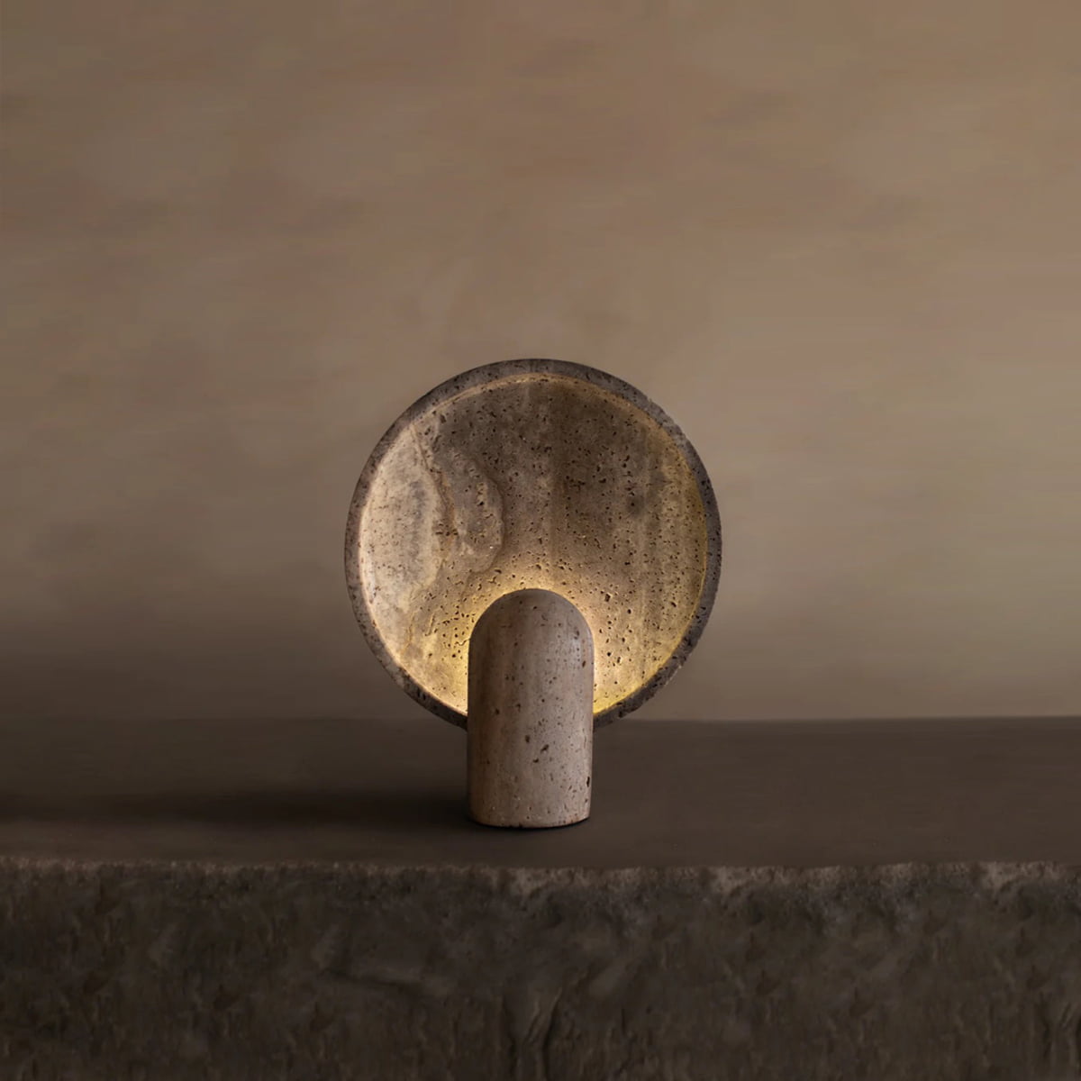 The Stella Lamp In Travertine features a round, travertine-inspired disc and cylindrical base, casting a soft, warm glow that enhances the serene ambiance of any space with its minimalist design and neutral tones.