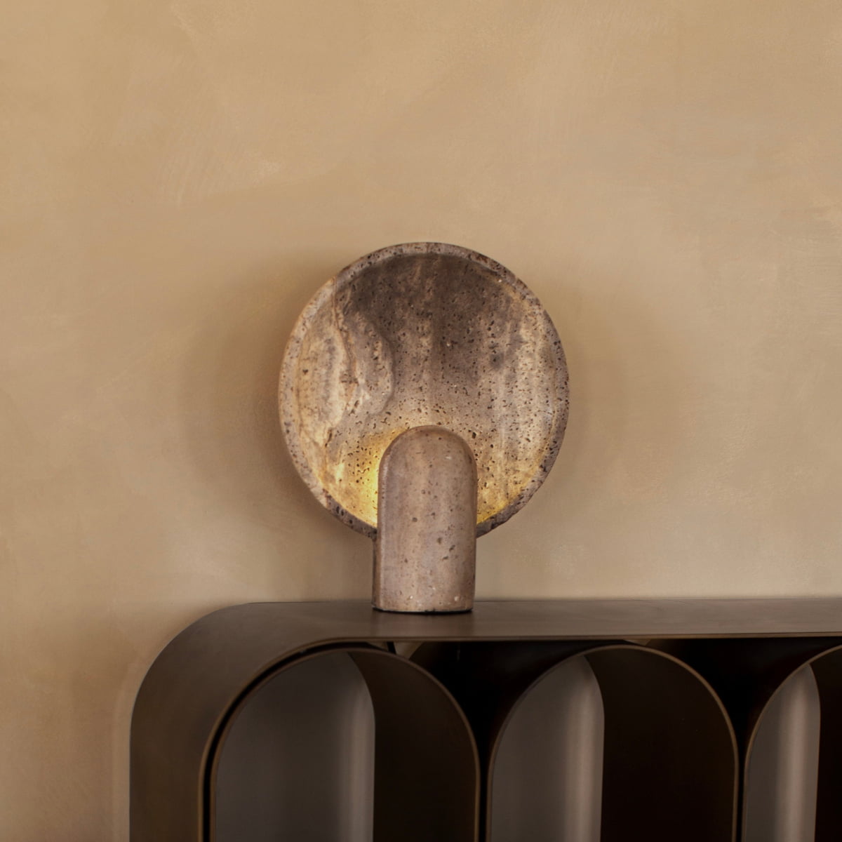 The Stella Lamp In Travertine, featuring a round textured stone design and cylindrical base, sits on a dark curved shelf against a beige wall, casting a soft glow that enhances the minimalist room’s calm and aesthetic appeal.