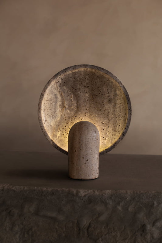 The Stella Lamp In Travertine features a minimalist cylindrical base and large circular plate, softly backlit to create a calming ambiance and warm glow against a neutral, textured backdrop.