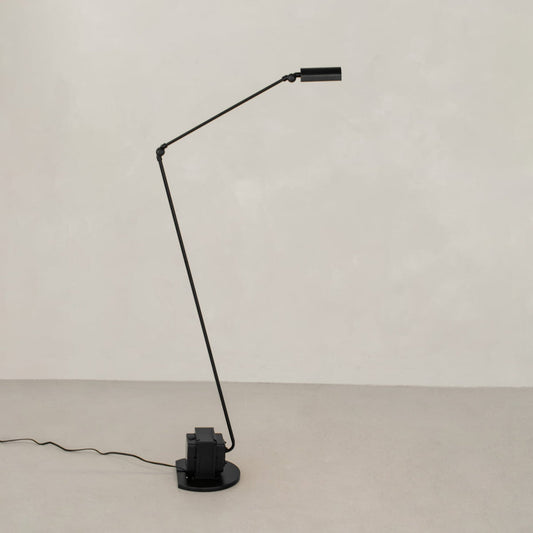 The black, modern Stem Lamp features a thin angled arm, cylindrical shade, and round base. Against a pale wall and floor, it offers adjustable height for versatile lighting, with the power cord lying neatly along the surface.