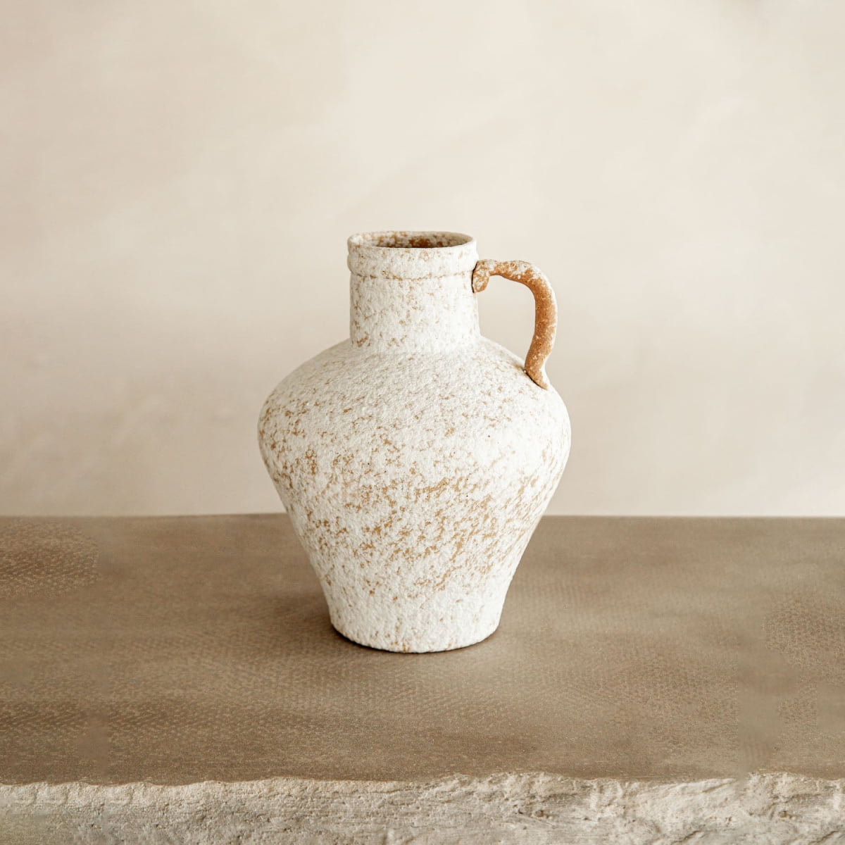 The Suna is a rustic, off-white ceramic jug with a narrow neck and single handle, resting on a textured beige surface—an inviting piece of artisan pottery ideal for farmhouse decor.
