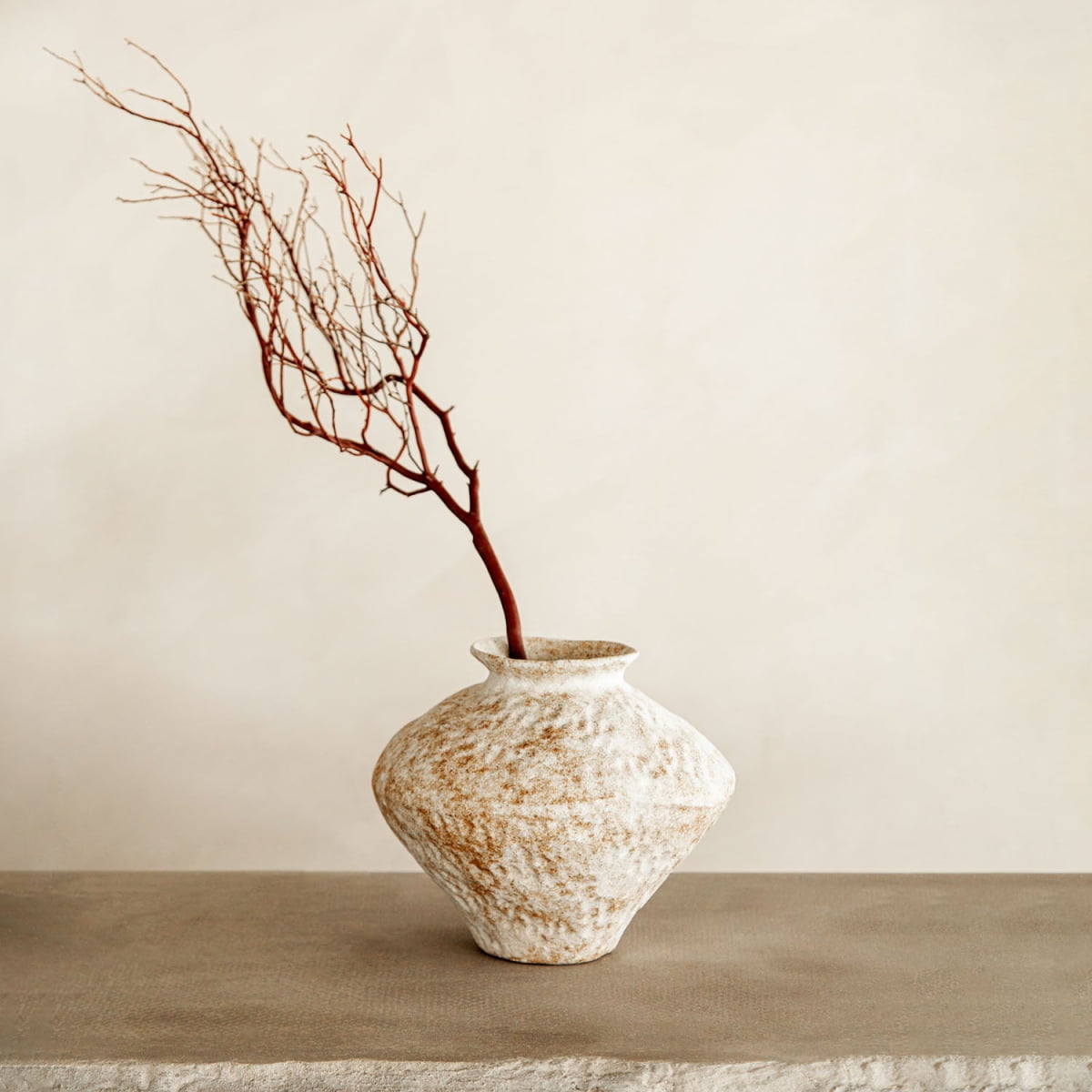 The Taru vase, a rustic textured white ceramic piece from artisan pottery, displays a single bare branch with reddish twigs on a neutral stone surface against a plain beige wall—ideal for refined home decor.