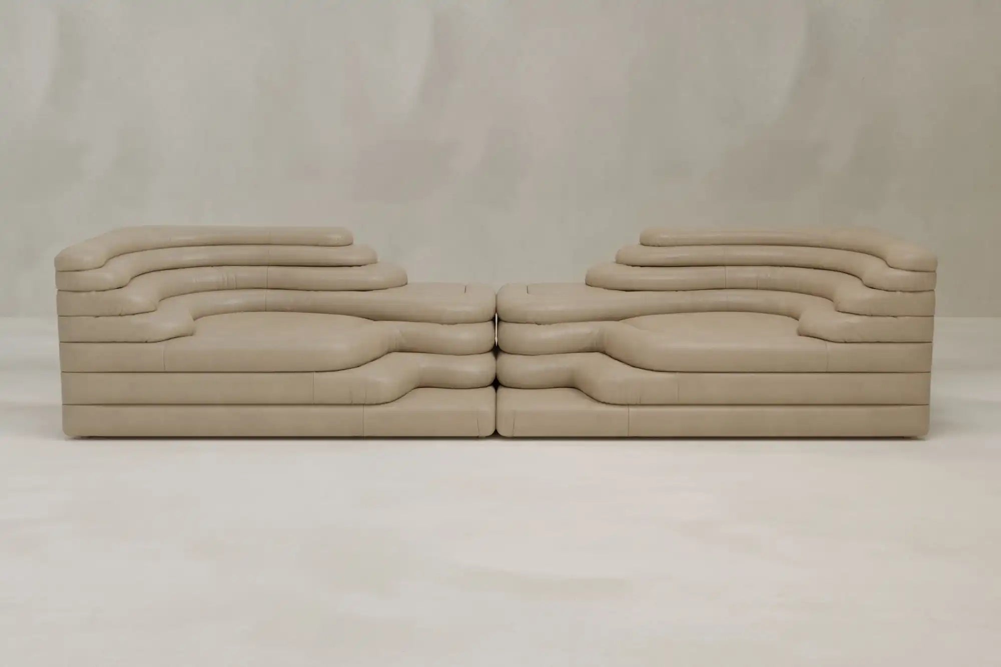 Two Cascata 2 Piece Sofa Rentals, with layered wavy forms and a sculptural beige design, face each other against a light background, adding modern sophistication to any space.