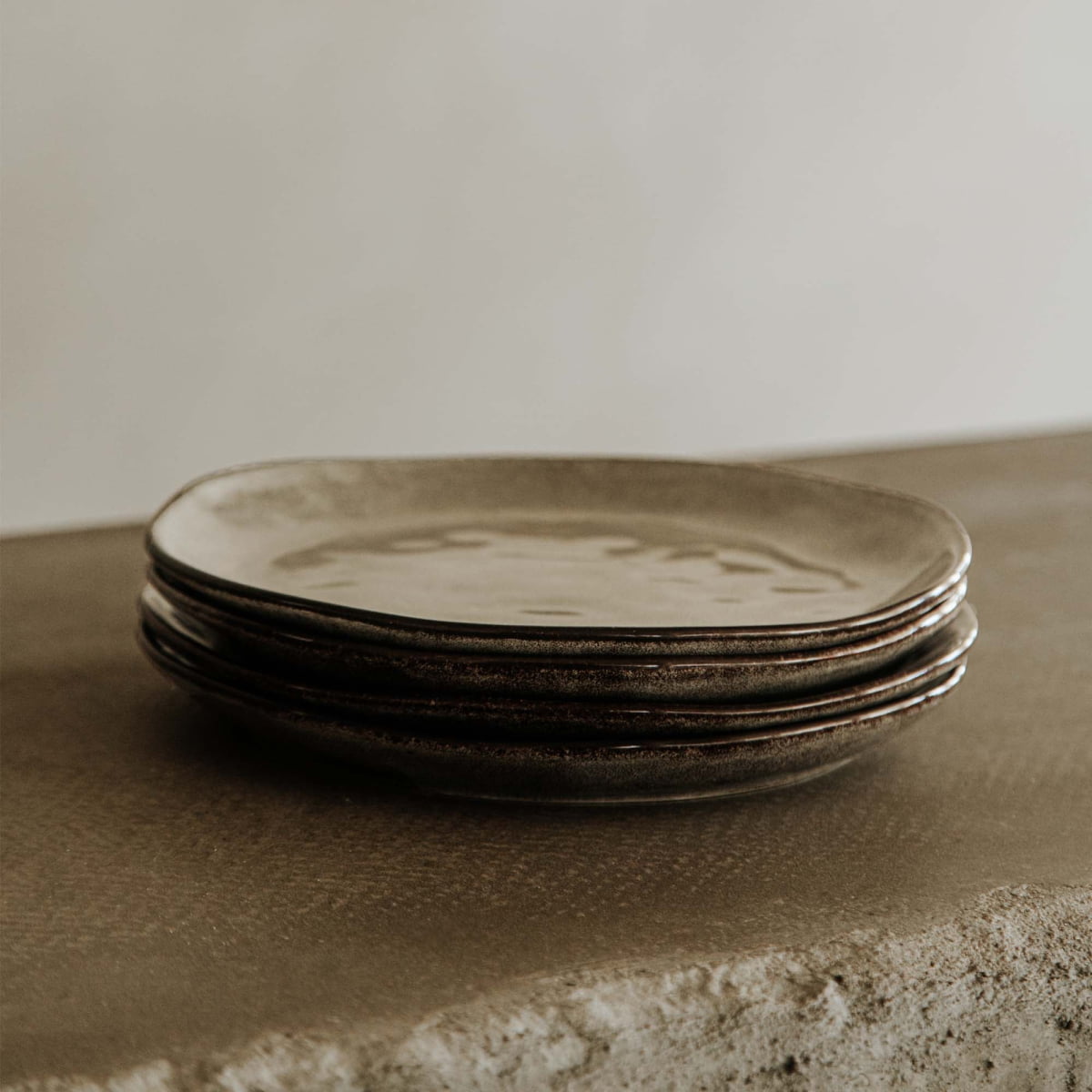 Four Tundra Glazed Dish Large plates with a rustic, matte ceramic finish are stacked on a textured stone surface, set against a soft, neutral backdrop.