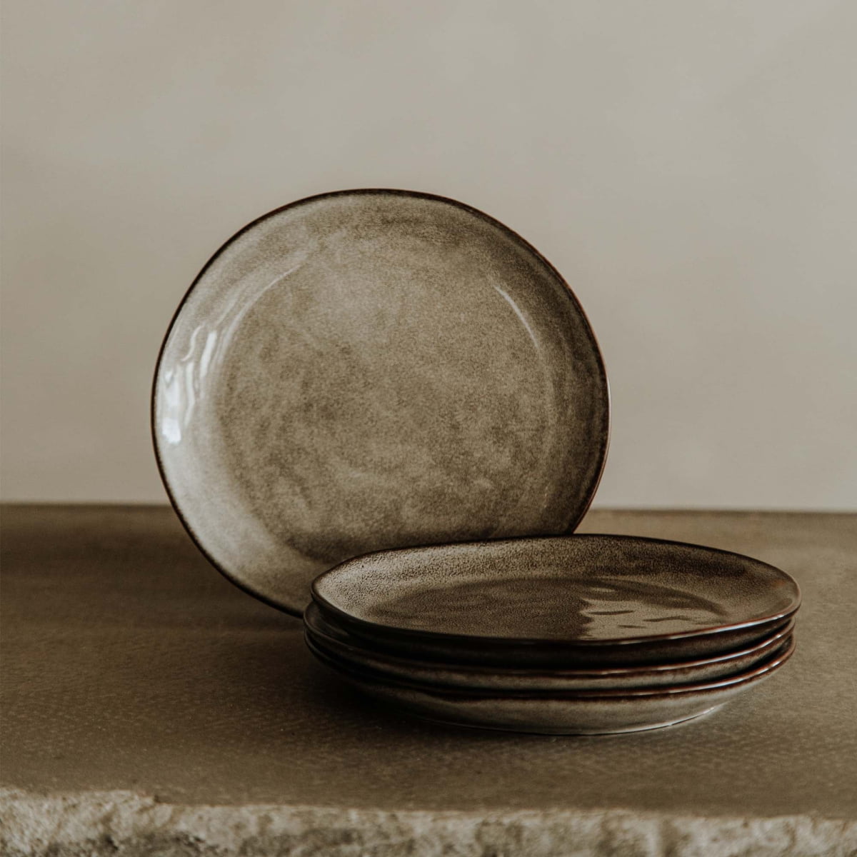 Four Tundra Glazed Dish Large ceramic plates with a rustic brown glaze are stacked on a textured stone surface, with one plate standing upright behind them against a neutral light beige background.