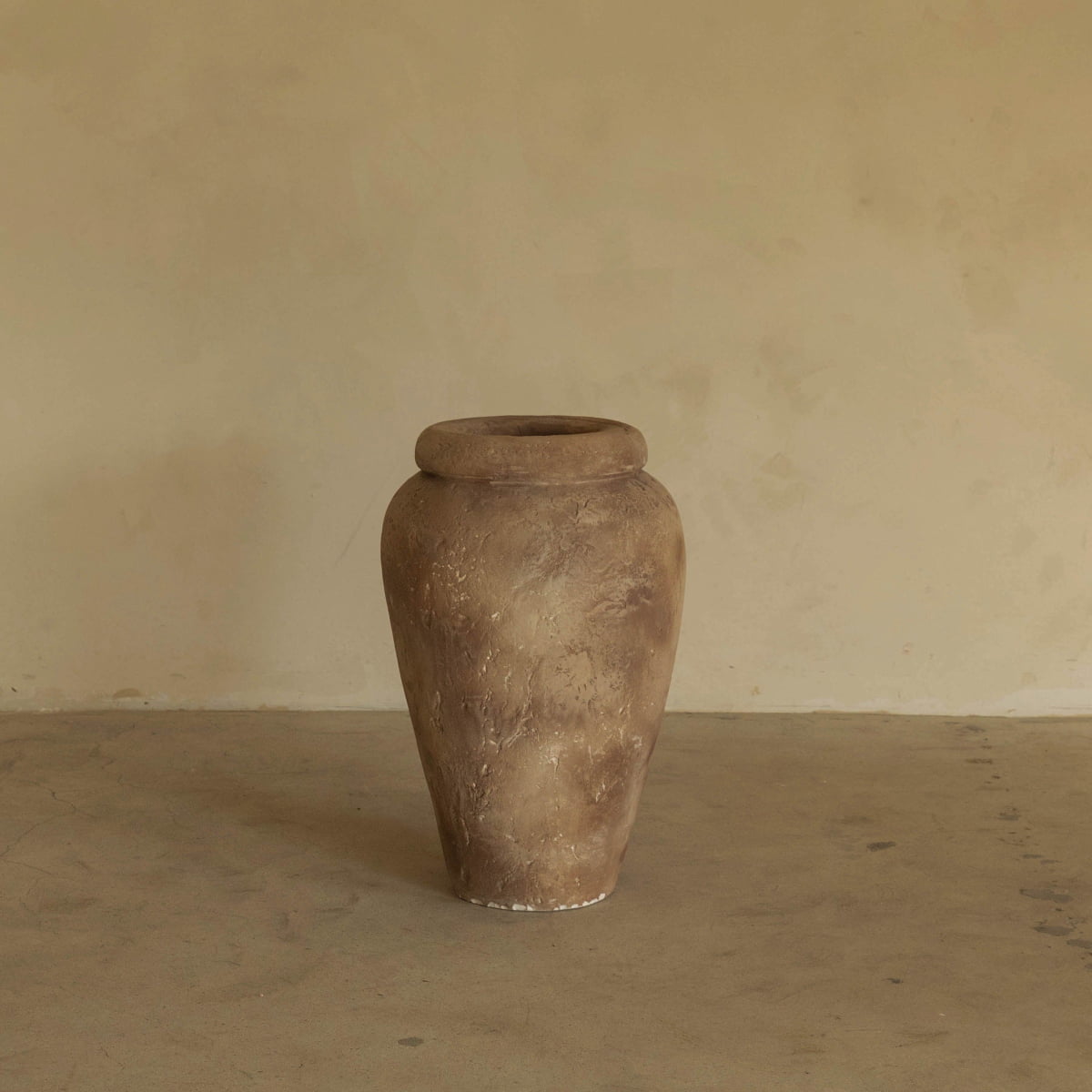 The Vase, a large hand-painted rustic clay piece with a wide rim made in Los Angeles, stands on a plain concrete floor against a beige textured wall.