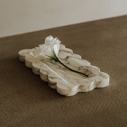 A single white flower rests on the Vera Marble Tray, shaped like a cloud, adding natural elegance to a brown textured surface.