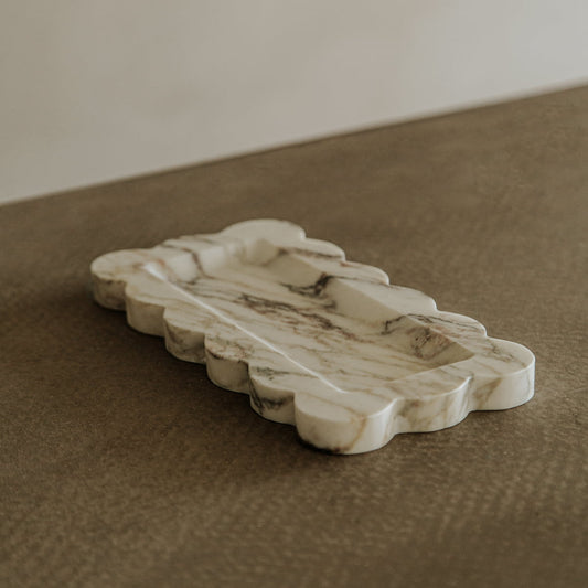The Vera Marble Tray features wavy, scalloped edges and beautiful beige and brown veining, creating a natural, elegant accent for any space. It sits stylishly on textured surfaces for versatile decor.