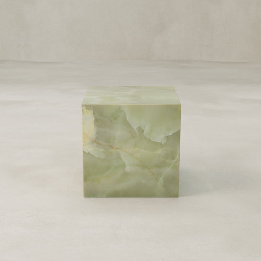 The Verdaine Onyx Plinth - Cube sits on a smooth, light beige surface against a matching backdrop, displaying natural stone veining and calming color shifts from pale green to cream.