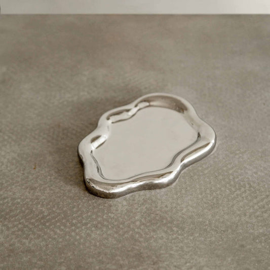 The Verve Tray, a small silver tray with wavy edges and a sculptural design, rests on a textured gray surface.