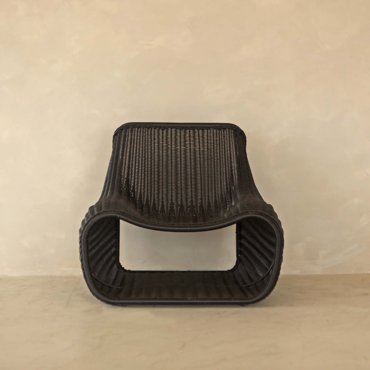 The Wicker Chair, featuring modern black PU rattan and a low, curved rectangular seat, is perfect for outdoor relaxation against a beige textured wall and floor.