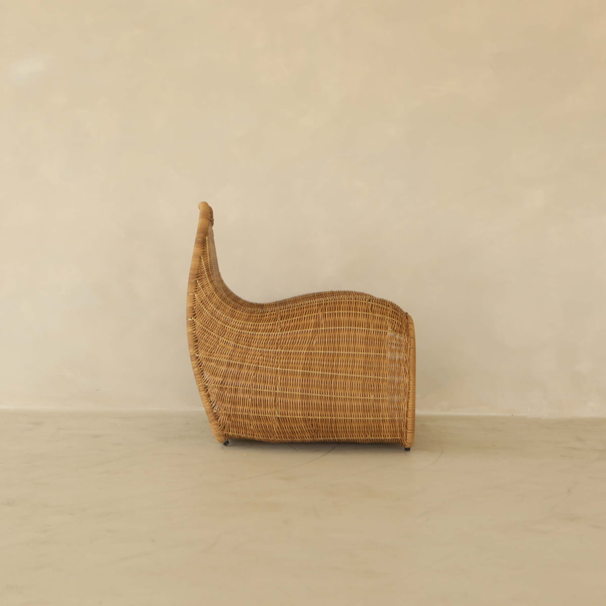 The Wicker Chair features a high, curved back and rounded base, placed sideways against a plain light wall and floor—perfect for relaxing outdoors.