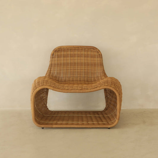 The Wicker Chair features a sculptural, curved design and woven PU rattan texture, set on a light floor against a beige wall—ideal for outdoor relaxation with its open, hollow base.