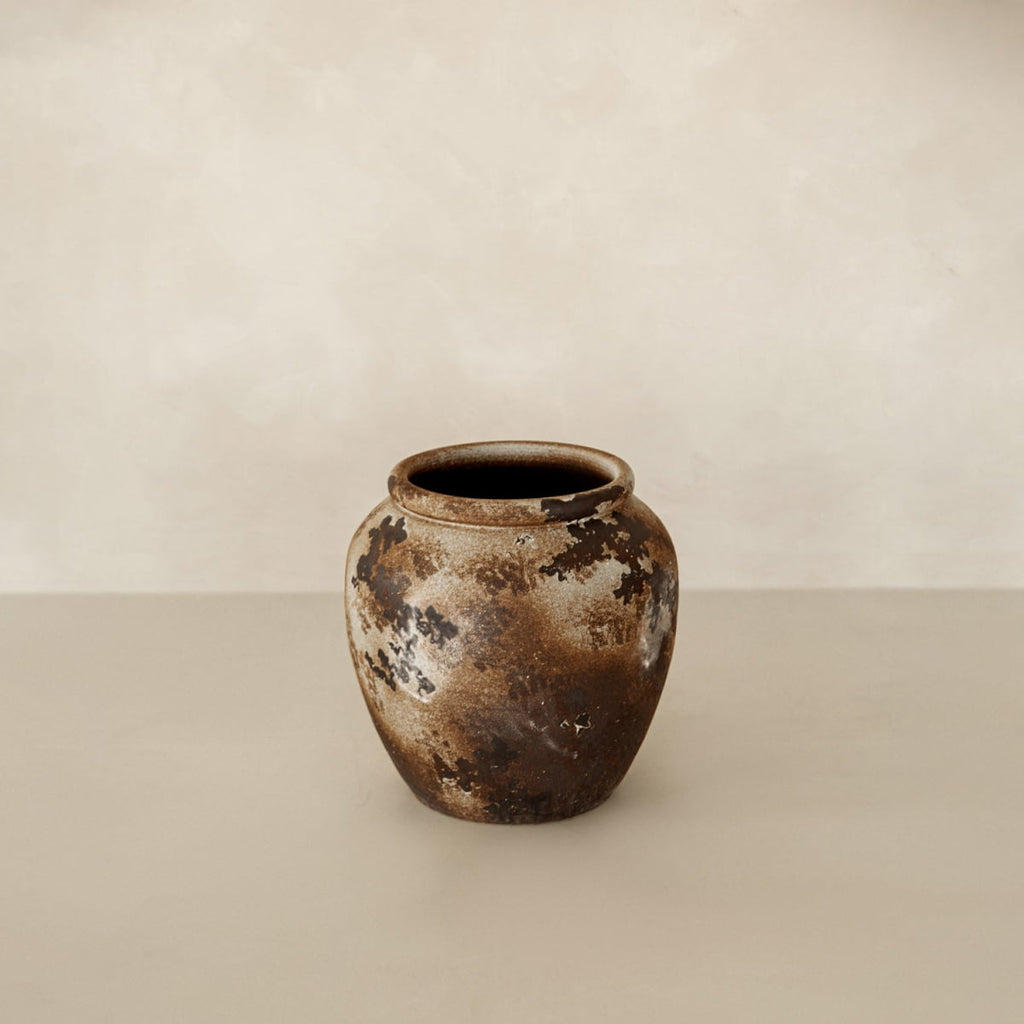 The Yogi vase, a rustic ceramic piece with a mottled brown and beige finish, sits on a neutral surface against a softly textured, pale background—showcasing handcrafted artisan charm.