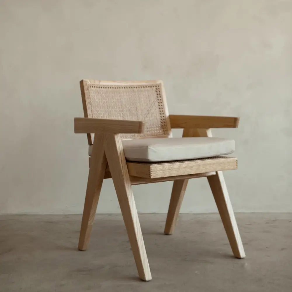 The Rivini Arm Chair features angled wooden legs, a hand-woven rattan cane backrest, and a light beige upholstered seat, shown on a concrete floor against a plain light wall.