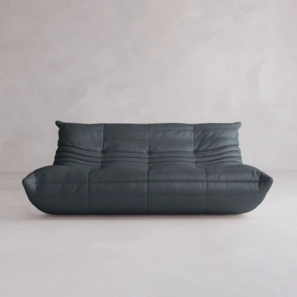 The Rolo Lounge Three Seater is a modern, armless sofa in dark gray with a low profile and plush tufted cushions, inspired by mid-century design, set against a plain light gray wall and floor.