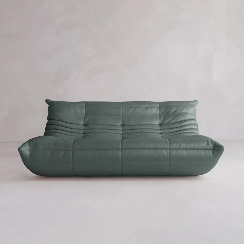 The Rolo Lounge Three Seater, a modern green leather sofa with deep tufting and mid-century inspiration, sits against a plain light-colored wall and floor.