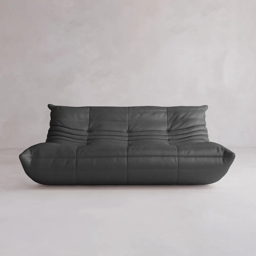 The Rolo Lounge Three Seater is a modern, low-profile black leather sofa with tufted cushions and mid-century inspired design, displayed against a plain light gray wall and floor.