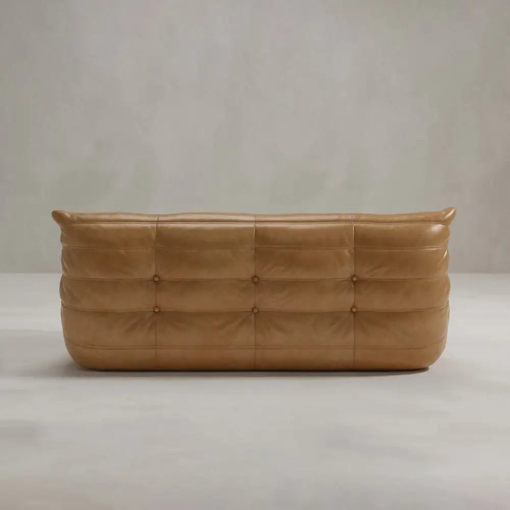 The Rolo Lounge Three Seater is a tan leather sofa with a low back, deep tufted cushioning, plush appearance, and subtle mid-century design elements, shown against a plain light-colored background.
