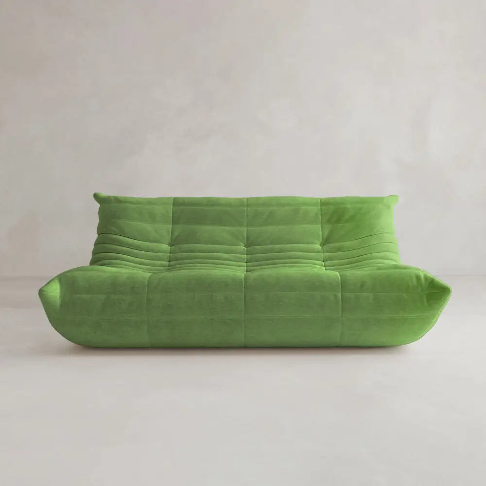 The Rolo Lounge Three Seater, a modern armless green sofa with low profile, tufted upholstery, and rounded edges, sits against a plain light-colored wall and floor, highlighting subtle mid-century design influences.