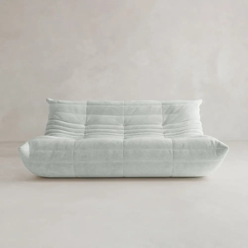 The Rolo Lounge Three Seater is a modern, light gray armless sofa with tufted, quilted details and a low backrest inspired by mid-century design, set against a plain, light-colored wall and floor.