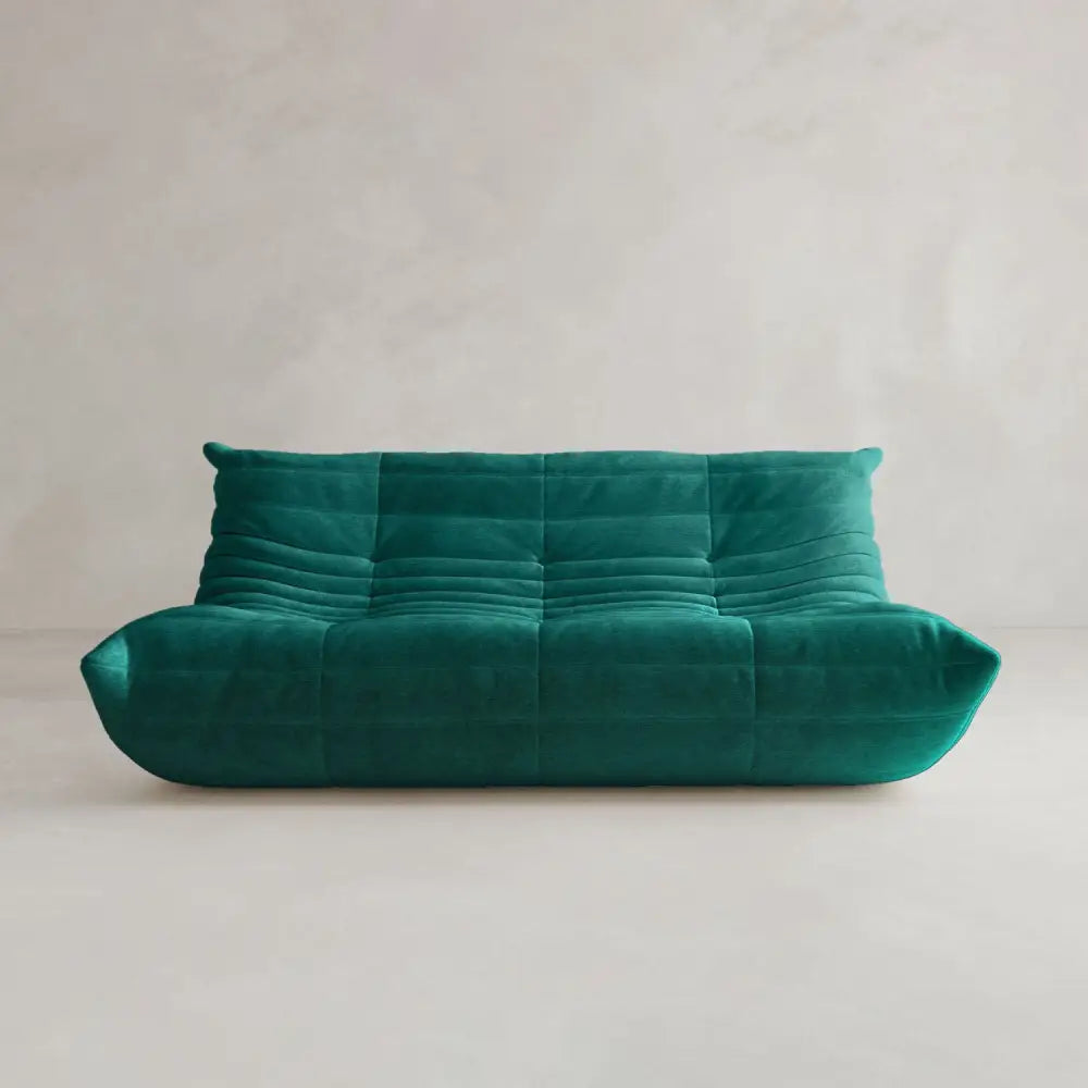 The Rolo Lounge Three Seater is a modern teal velvet sofa with a low profile, tufted seat and back, and soft rounded edges that showcase mid-century design, set against a plain light-colored wall and floor.