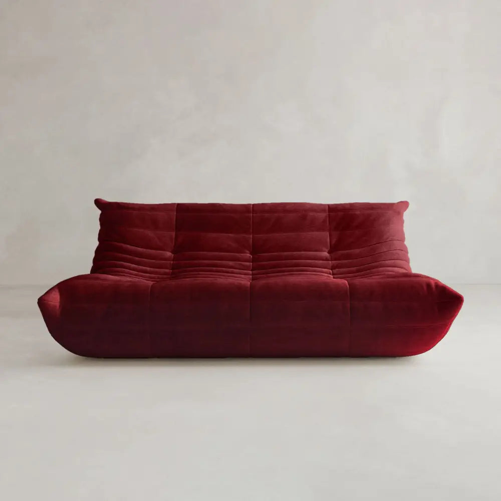 The Rolo Lounge Three Seater, a deep red velvet sofa with tufted cushions and a minimalist mid-century design, features a low profile and is set against a light gray textured background.