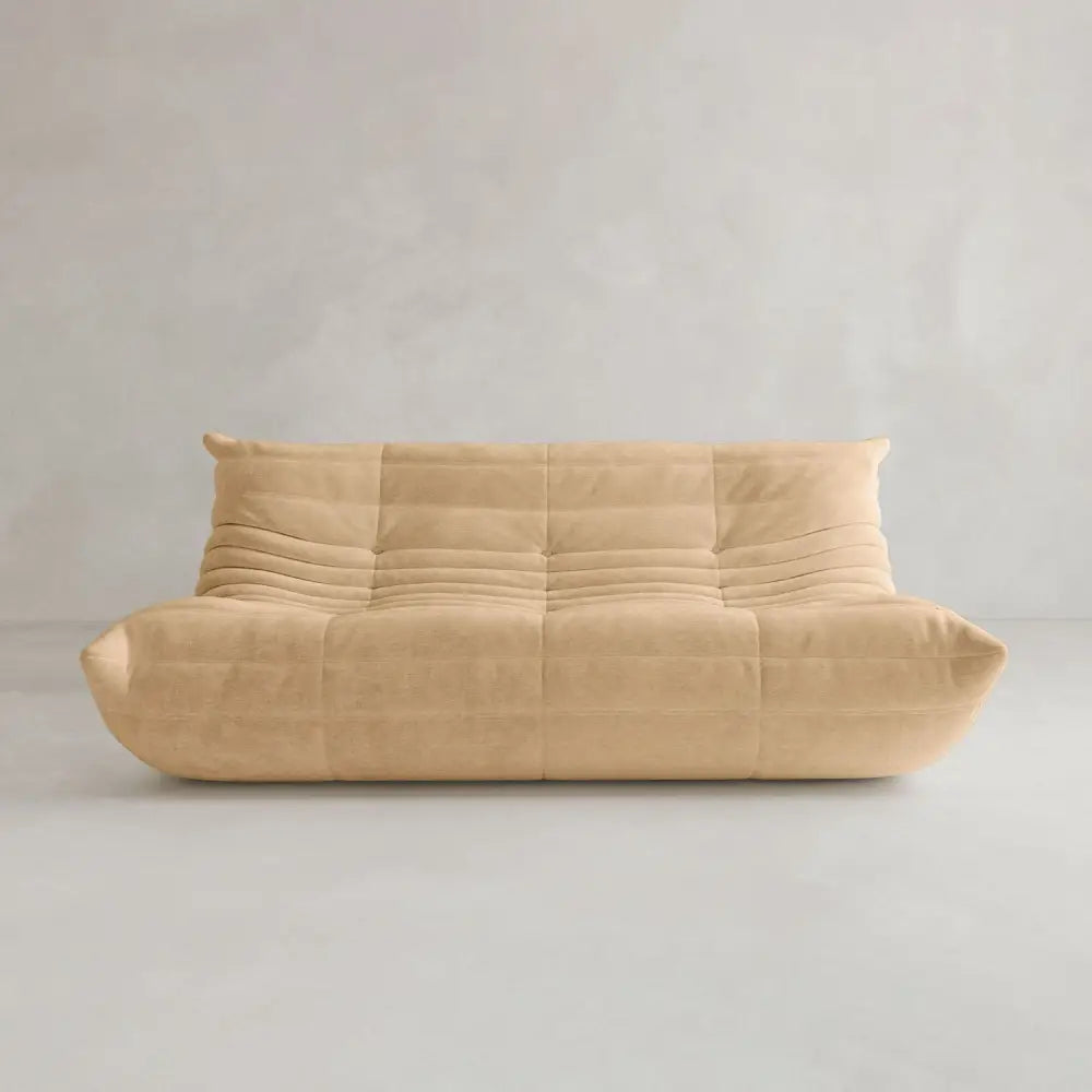 The Rolo Lounge Three Seater, a modern armless beige sofa with a quilted cushioned design inspired by mid-century style, sits against a plain light gray wall and floor.