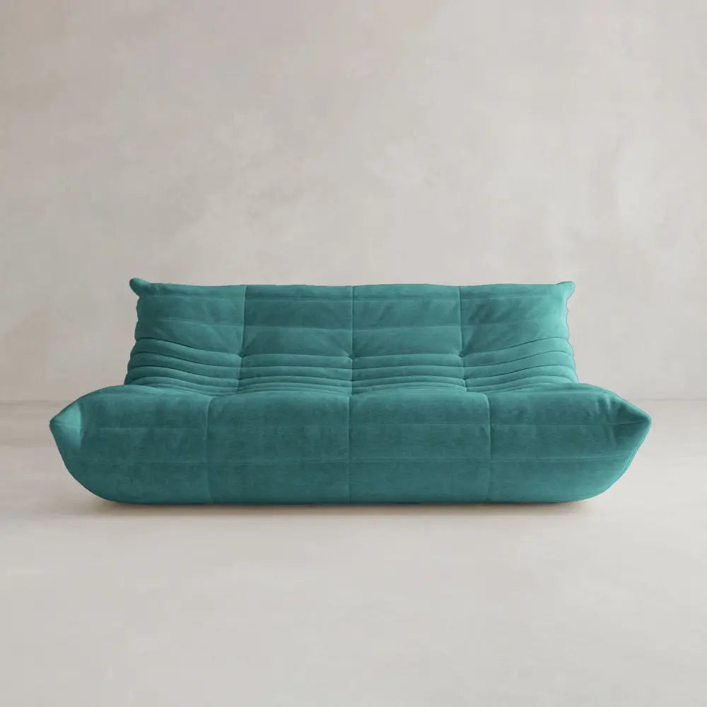 The Rolo Lounge Three Seater, a modern teal sofa with plush, quilted upholstery and a low backrest inspired by mid-century design, sits against a light-colored wall in a minimalistic room.