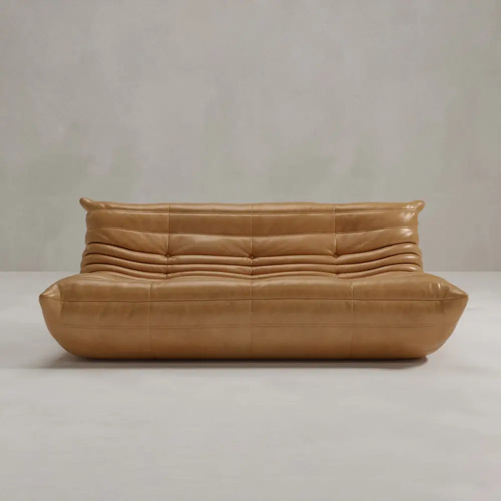 The Rolo Lounge Three Seater features tan leather upholstery with a smooth, tufted design and horizontal pleats, inspired by mid-century style. Its modern, low-profile shape has rounded edges, no visible legs, and is set against a light gray background.