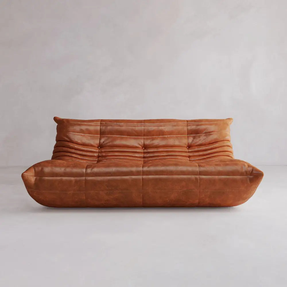 The Rolo Lounge Three Seater, a modern brown leather sofa with plush wrinkled cushions and a low profile, sits against a plain light gray wall and floor. Its minimalist design draws on mid-century style with no visible legs or armrests.