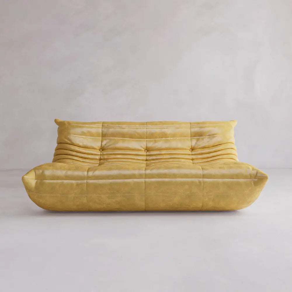 The Rolo Lounge Three Seater, featuring mustard yellow leather and plush, horizontally tufted cushions, brings mid-century style to life against a simple light gray wall and floor.