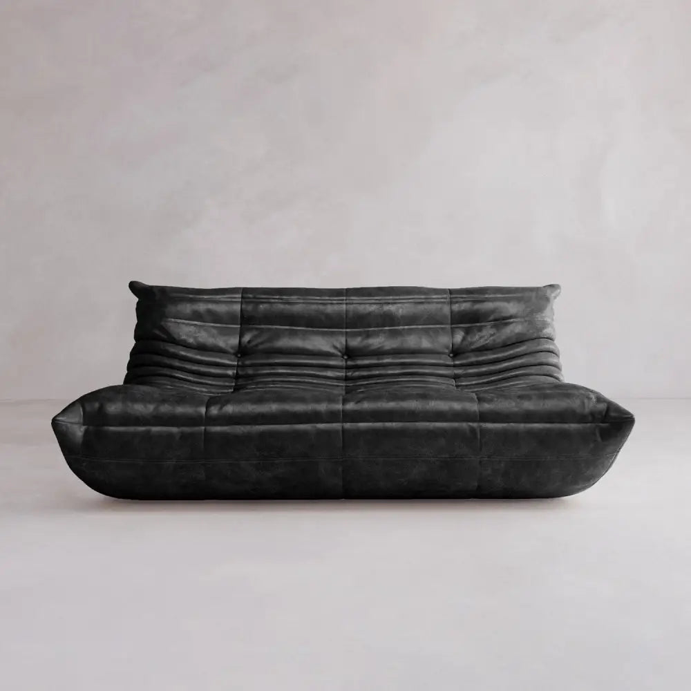 The Rolo Lounge Three Seater is a modern black leather sofa with a low back, deep seat, soft gathered cushions, and tufted details—echoing mid-century design—set against a plain light gray wall and floor.