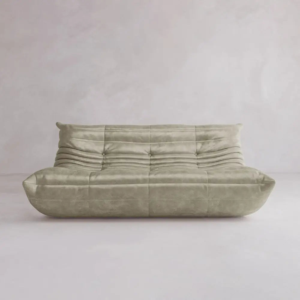 The Rolo Lounge Three Seater—a modern, light beige sofa with a low backrest, deep seat, and soft gathered upholstery inspired by mid-century design—sits against a plain, light-colored wall and floor.