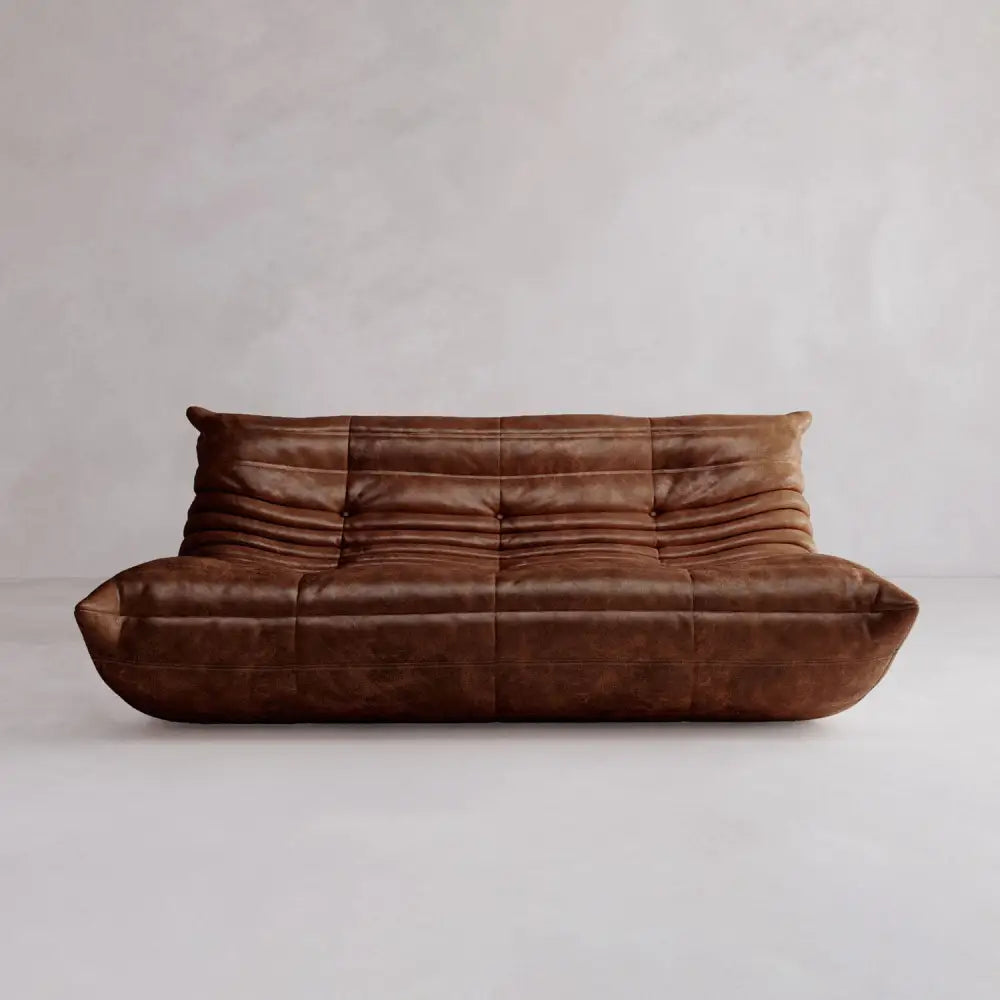 The Rolo Lounge Three Seater is a brown leather sofa featuring a modern, low-profile silhouette and soft, rounded edges inspired by mid-century design, shown against a light, plain background.
