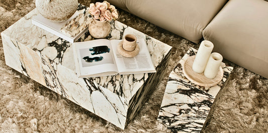 Modern luxury coffee table in striking marble with bold veining, styled with books, ceramics, and flowers on a textured rug beside a neutral sofa.