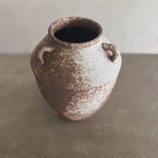 The Yuki vase is a rustic, brown and white ceramic piece with two small handles near the top, displayed on a neutral surface against a blurred background.