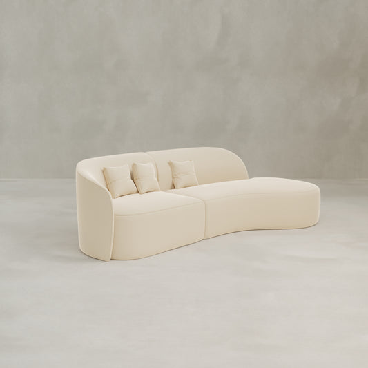 The Marlow Curved Modular Three Seater Sofa features modern cream-colored upholstery and comes with three matching throw pillows, shown in an empty room with light gray walls and a smooth, pale floor.