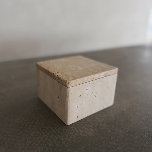 The Celeste Vault with Lid, a small square beige stone box, rests on a dark textured surface with a softly blurred neutral background.