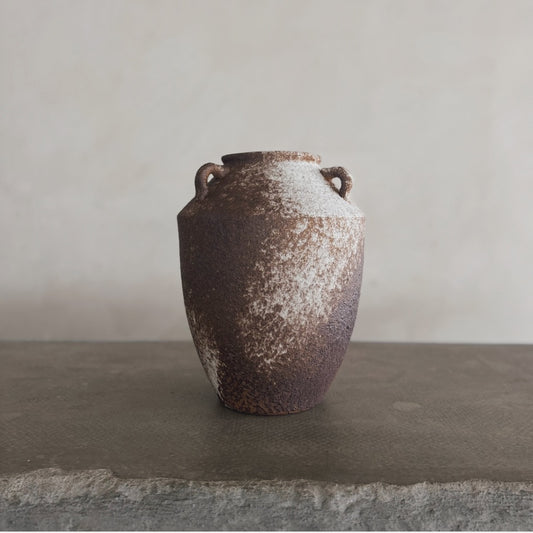 The Yuki is a rustic brown and white ceramic vase with two small side handles, displayed on a textured stone surface against a plain, light-colored background.