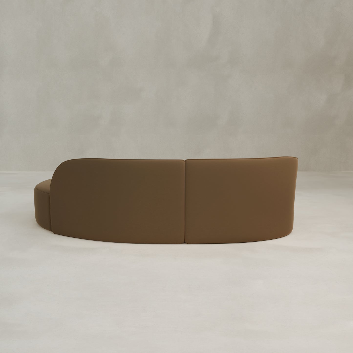 The Marlow Curved Modular Three Seater Sofa, upholstered in brown velvet, is placed against a light grey textured wall on a smooth white floor and viewed from behind.