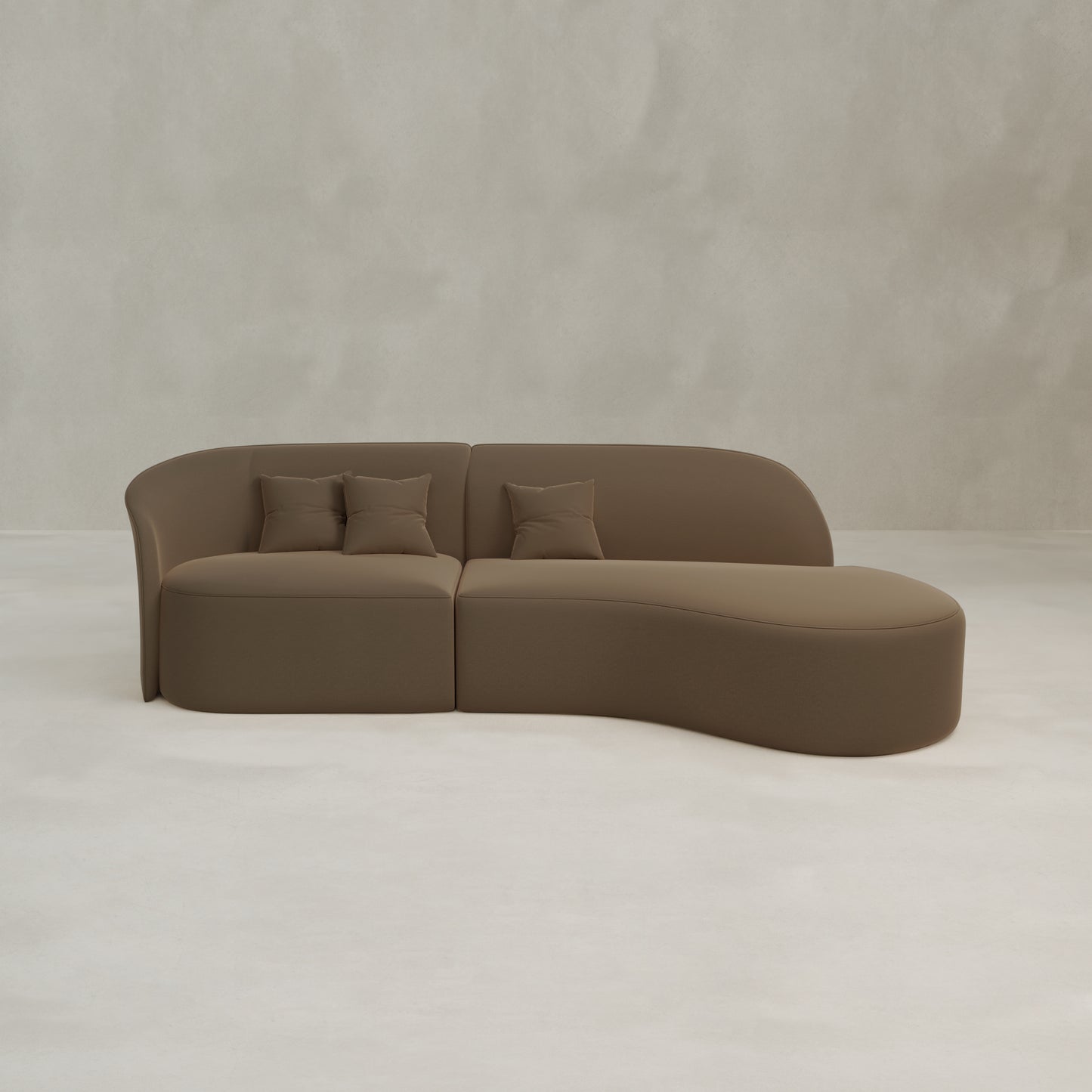 The Marlow Curved Modular Three Seater Sofa in brown velvet features a luxurious curved design, chaise lounge, two seat cushions, and three matching pillows, set against a light gray textured wall and floor.