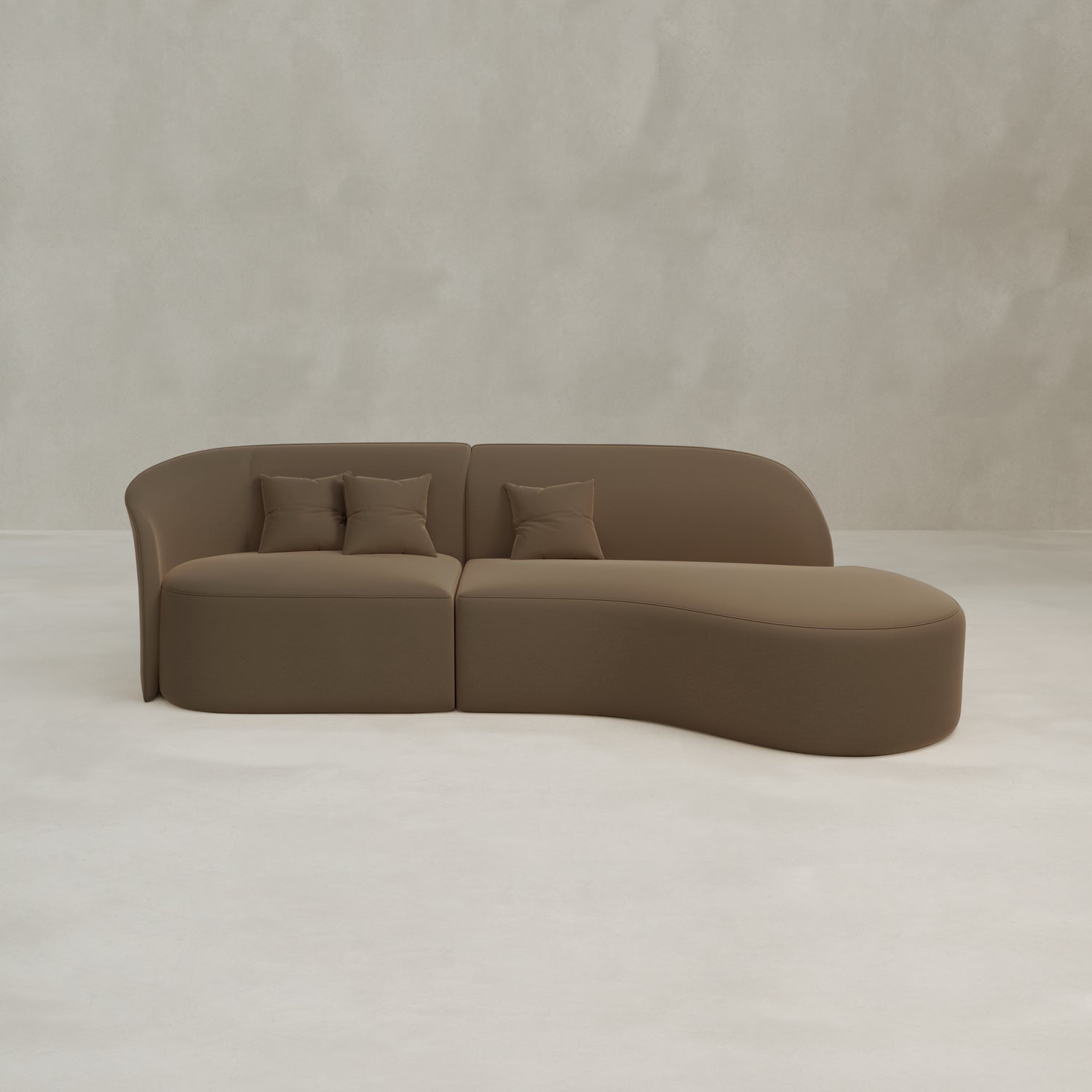The Marlow Curved Modular Three Seater Sofa in brown velvet features a luxurious curved design, chaise lounge, two seat cushions, and three matching pillows, set against a light gray textured wall and floor.